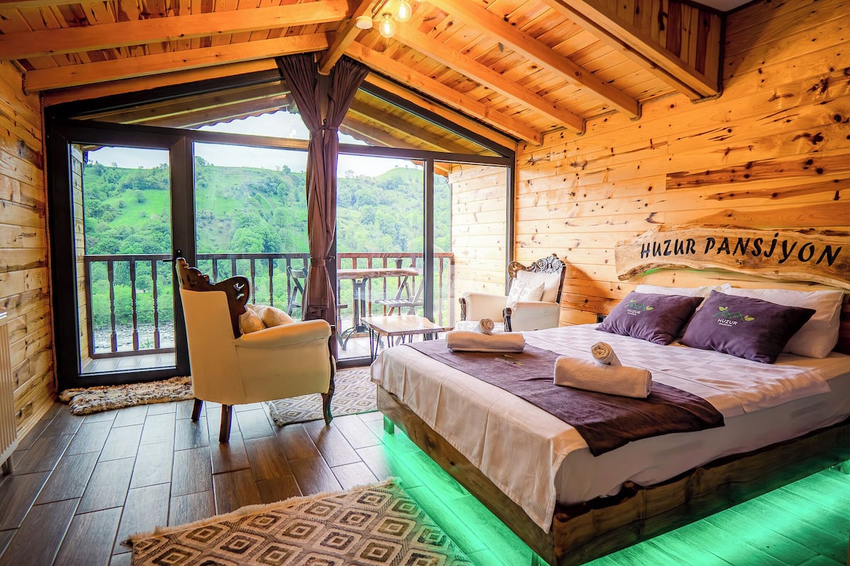 A wooden cabin room with a bed, a chair, a table, and a balcony with a view of green hills.
