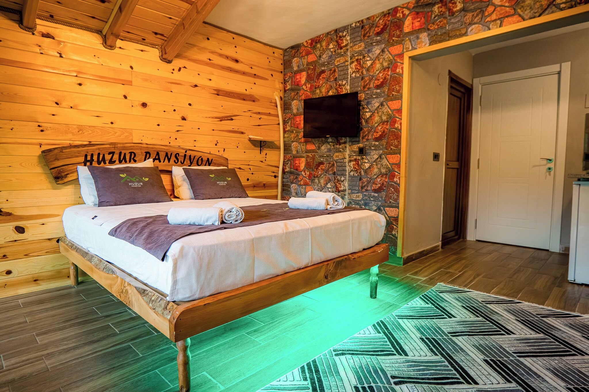 Un letto di legno con una testiera che recita 'Huzur Pansiyon', una parete con effetto pietra, un televisore a schermo piatto e un tappeto verde a motivi.