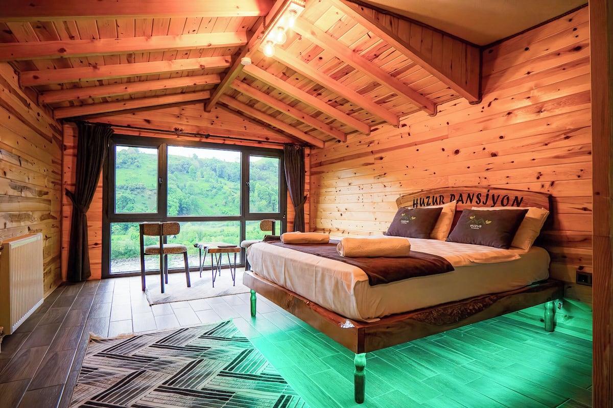 A wooden cabin bedroom with a bed, a chair, a view of greenery, and a window.