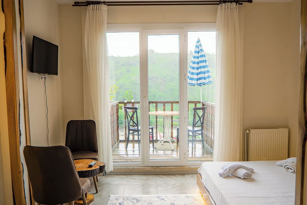 A hotel room with a bed, a desk with a chair, a TV, a balcony with a table and chairs, and a view of greenery.