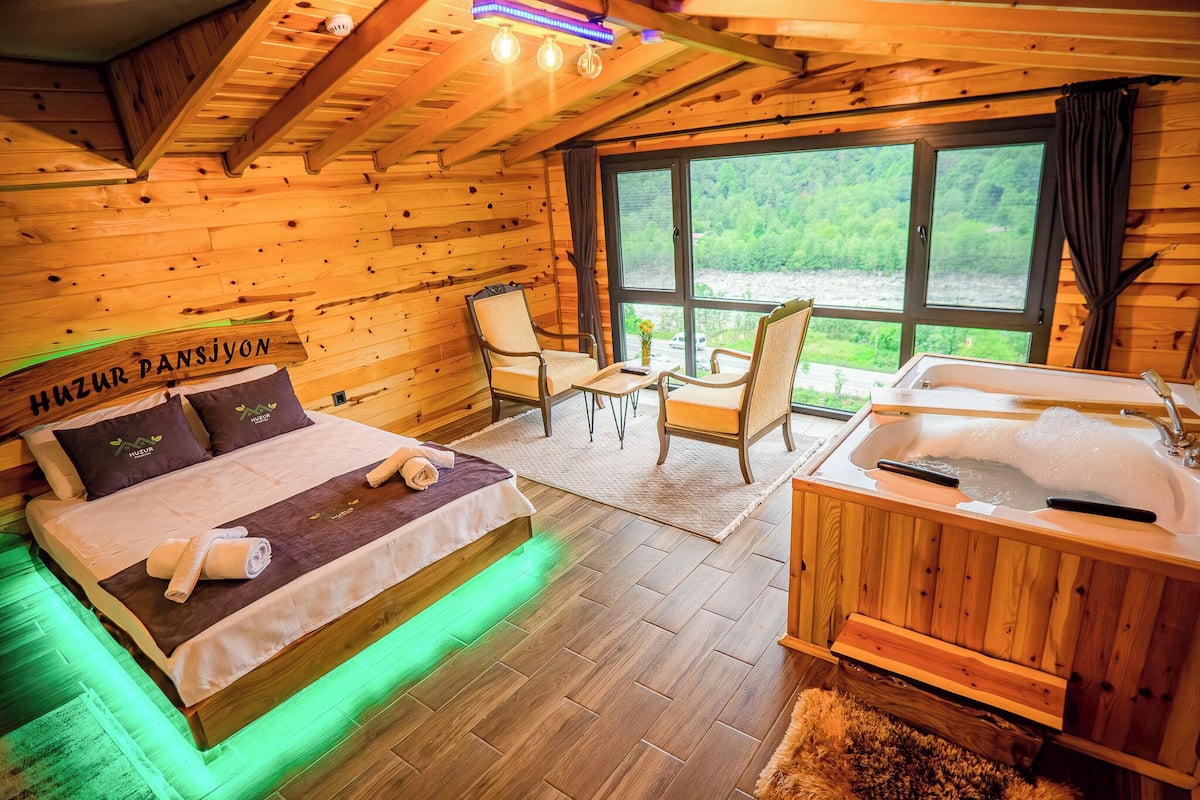 A wooden cabin room with a bed, two chairs, a small table, a bathtub, and a large window with a view of greenery.