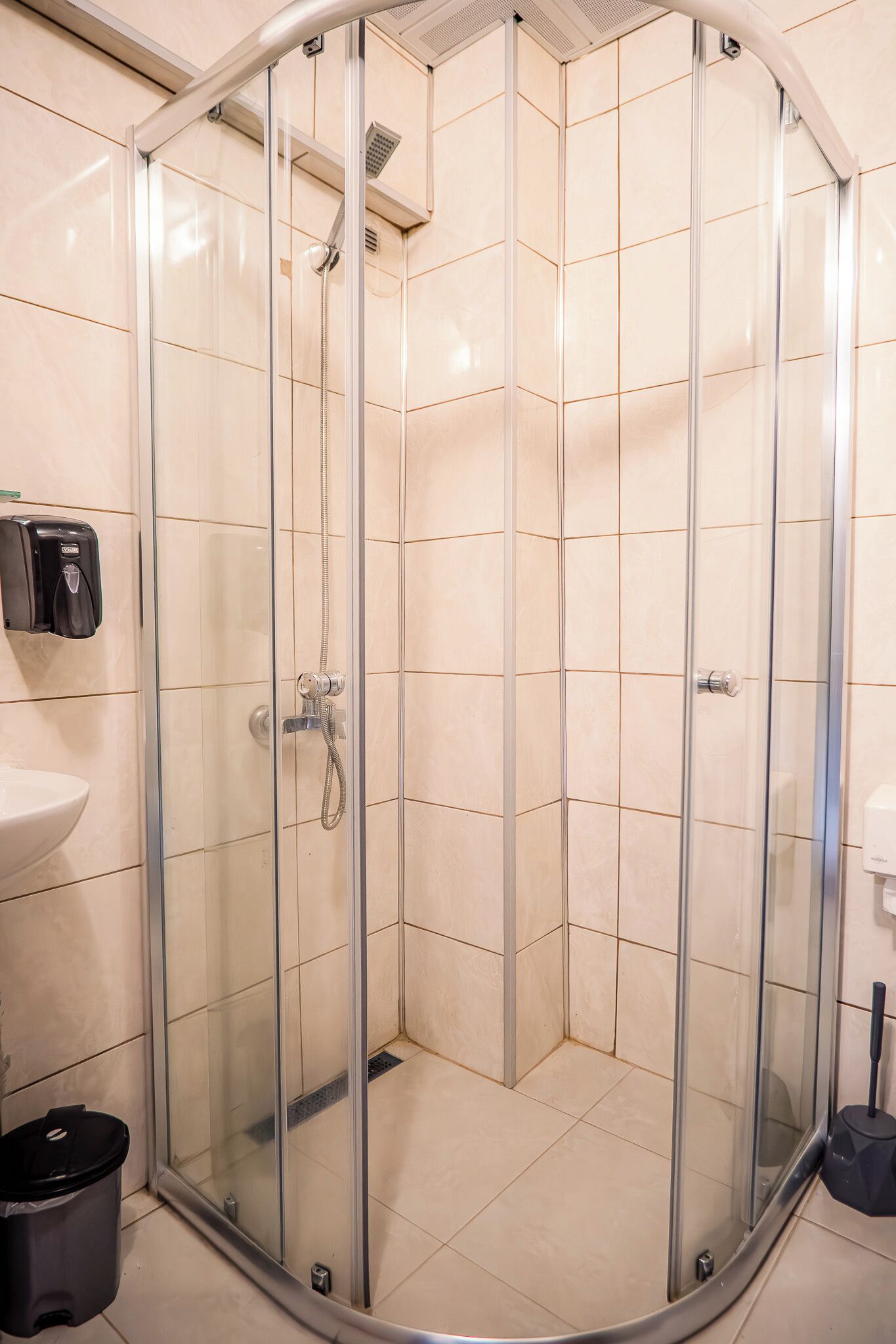 A glass-enclosed shower stall with a handheld showerhead, a wall-mounted soap dispenser, and a trash bin.