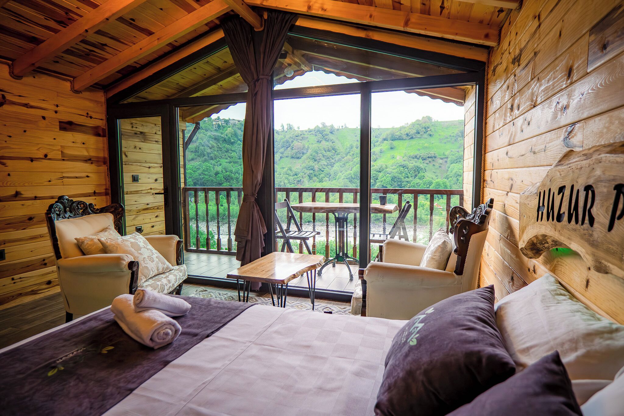 A wooden cabin room with a bed, two chairs, a table, and a large window offering a view of a forested hillside.