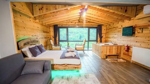 Interior de una cabaña de madera con cama, sofá, mesa de comedor y vistas amplias a través de grandes ventanas.