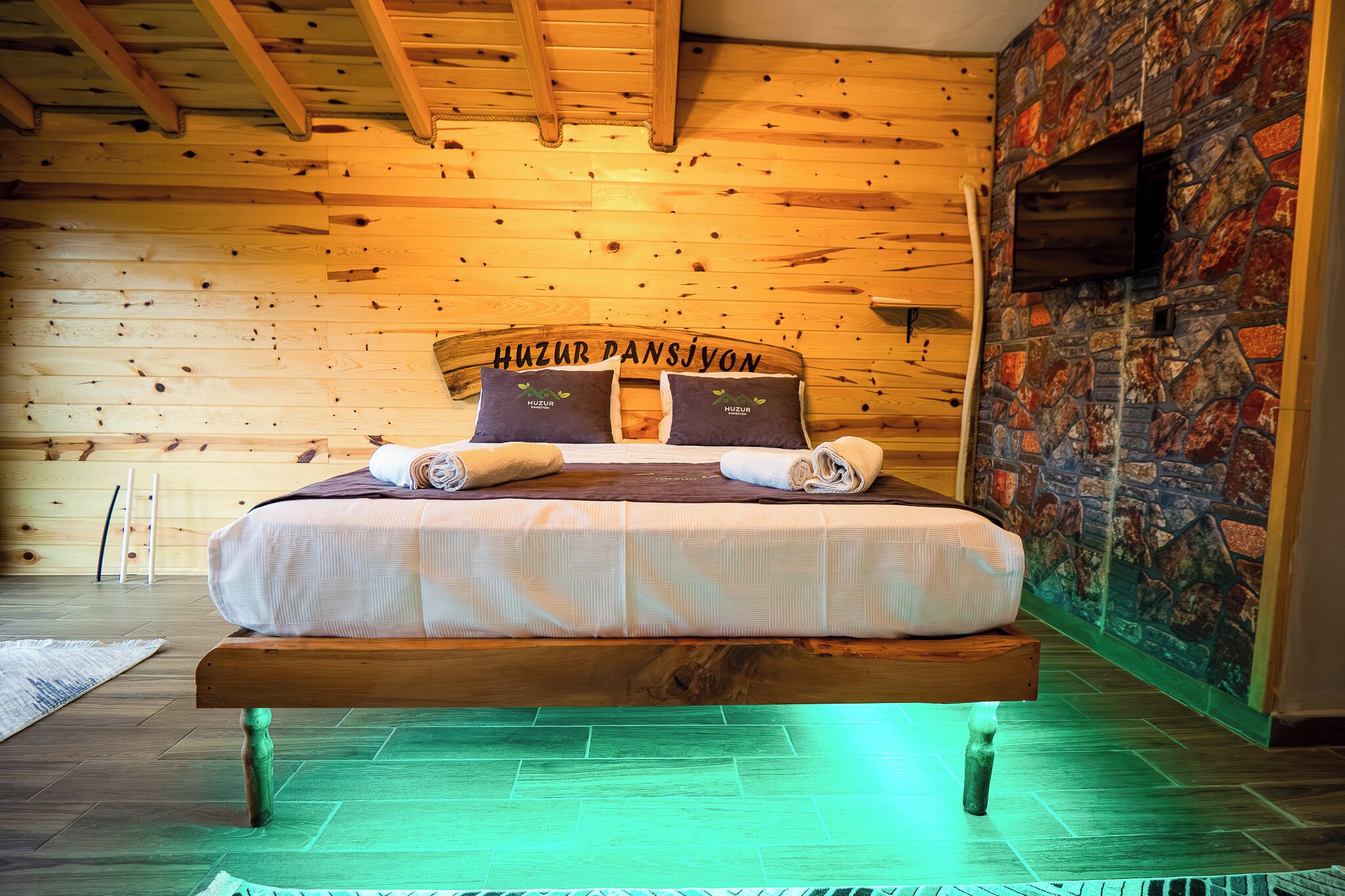 Een houten bed met hoofdeinde waarop 'Huzur Dansiyon' staat, in een kamer met houten wanden en een accentmuur van steen.