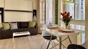 A modern living room with a sofa, coffee table, dining table, and chairs. There is a window with a view of buildings outside. - Ferienwohnung in Sellin mit Balkon (Sellin)