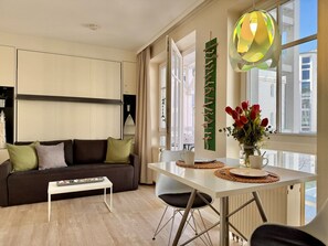 A modern living room with a sofa, coffee table, dining table, and chairs. There is a window with a view of buildings outside. - Ferienwohnung in Sellin mit Balkon (Sellin)