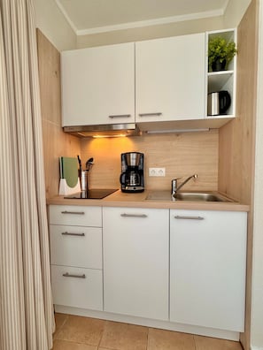 A compact kitchen with white cabinets, a coffee maker, a sink, and a shelf with a plant. - Ferienwohnung in Sellin mit Balkon (Sellin)