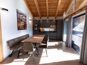 A modern interior with a wooden dining table, benches, and a kitchen area with a large window. - Ski-in/ski-out Chalet With Beautiful View (Sankt Lambrecht)