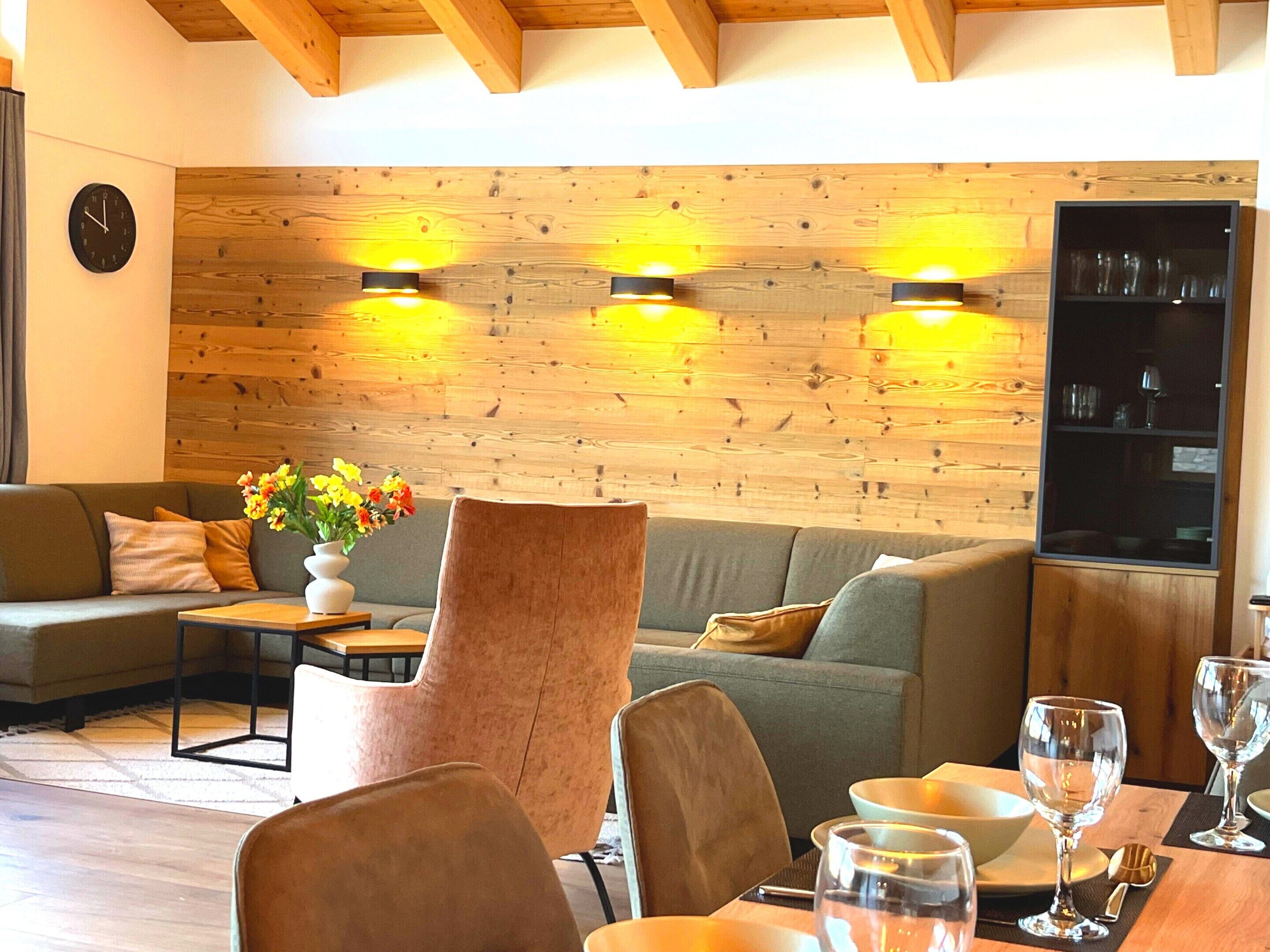 Un salon chaleureux avec un mur en bois, un canapé, un fauteuil, une table à manger avec des verres et une horloge au mur.