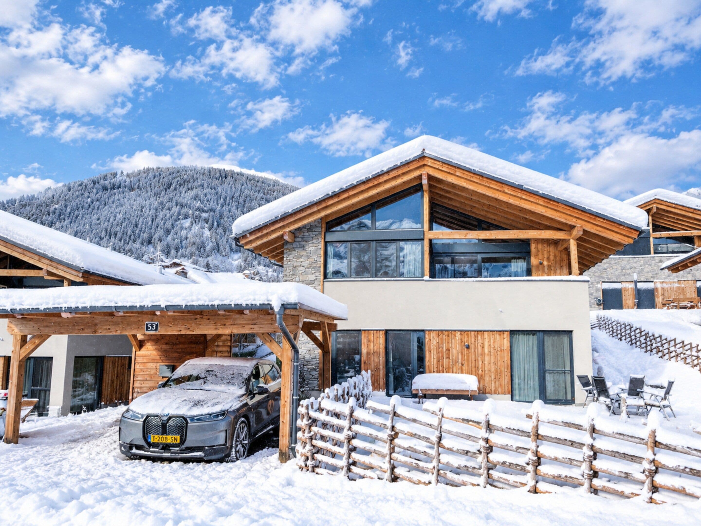 A modern chalet with a wooden exterior, a car parked in front, and a snow-covered mountain in the background.