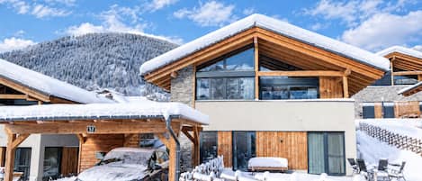 Un moderno chalet con fachada de madera, un coche estacionado adelante y una montaña nevada al fondo.
