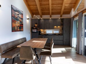 A modern kitchen with wooden beams, a dining area with a wooden table and benches, and a large window with curtains. - Ski-in/ski-out Chalet With Beautiful View (Sankt Lambrecht)