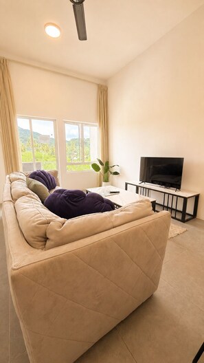 A living room with a beige sofa, a television on a stand, and a window with curtains. - Spacious 3-bedroom Mountain View Apartment in Langkawi (Langkawi)
