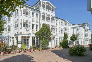 Ein weißes Gebäude mit mehreren Balkonen und einer Pflasterfläche, bepflanzt mit Blumen und mit Schildern.
