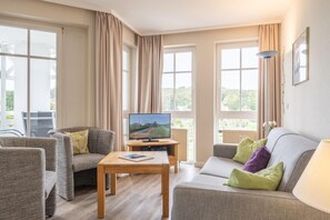 Ein Wohnzimmer mit einer Couch, Sesseln, einem Couchtisch, einem Fernseher und großen Fenstern.