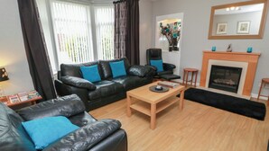 A living room with a black leather sofa, a wooden coffee table, a fireplace, and a large window with curtains. - 1 St. Catherines Cottages (Perth)