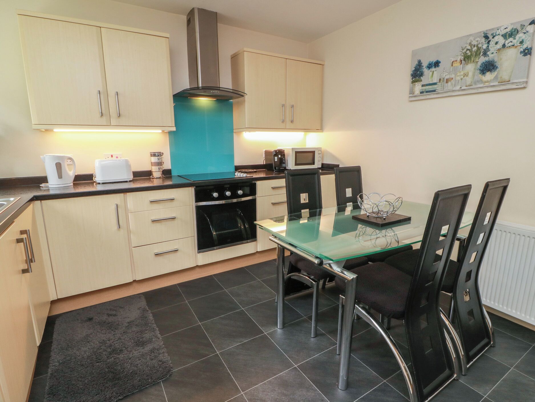 A compact kitchen with a stove, microwave, and cabinets, a glass dining table with chairs, and a painting on the wall.