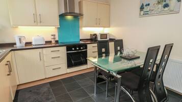 A compact kitchen with a stove, microwave, and cabinets, a glass dining table with chairs, and a painting on the wall.