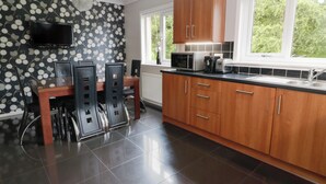 A modern kitchen with wooden cabinets, a dining table with chairs, a flat-screen TV mounted on the wall, and a window providing natural light. - Cliff View (Perth)