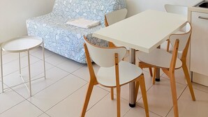 A modern, minimalist living space with a sofa, a dining table with chairs, a small kitchen area, and a window with blinds. - St Cyprien, a sunny interlude by the water (Saint-Cyprien)