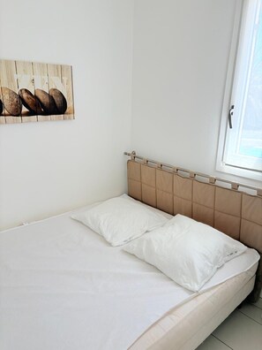 Uma cama de solteiro com cabeceira, janela com vista e um quadro fixado na parede.
