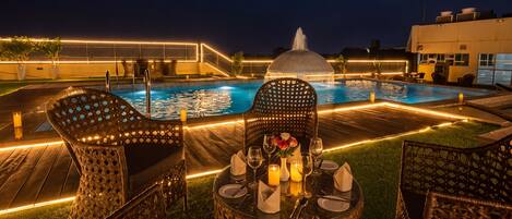 Zona de piscina con mobiliario de mimbre, una mesa lista para comer y una fuente al fondo.