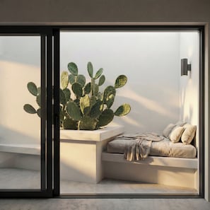 A modern room with a sliding glass door, a cactus plant, a cushioned bench, and a wall-mounted light fixture. - Serenity Villa, Anzo Villas (Thira - Santorini)
