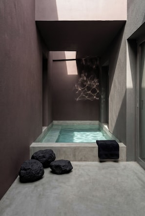 A modern spa area with a rectangular hot tub, dark textured rocks, and a black towel on a platform. - Serenity Villa, Anzo Villas (Thira - Santorini)
