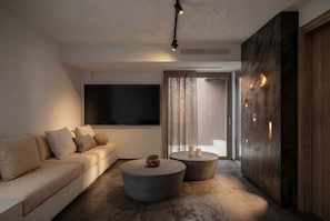 A modern living room with a sofa, two round coffee tables, a flat-screen TV, and a large window with curtains. - Serenity Villa, Anzo Villas (Thira - Santorini)