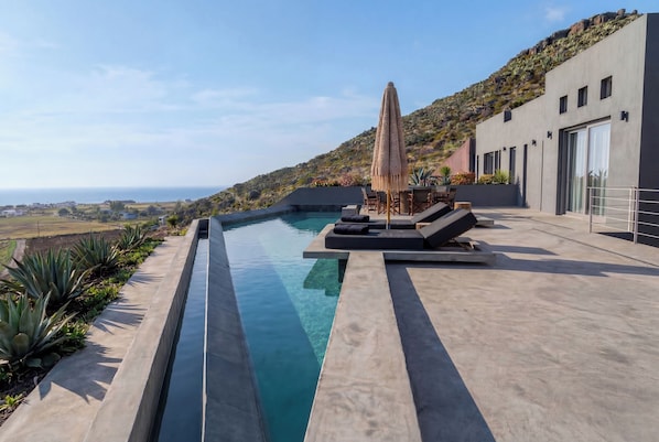 Ein moderner Poolbereich im Freien mit freiem Blick auf den Ozean, eine Lounge mit Sitzgelegenheiten und eine Hügelkulisse.