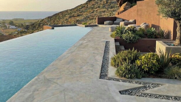 Una piscina infinita moderna con vistas claras al océano, un área ajardinada con plantas y un paisaje montañoso de fondo.