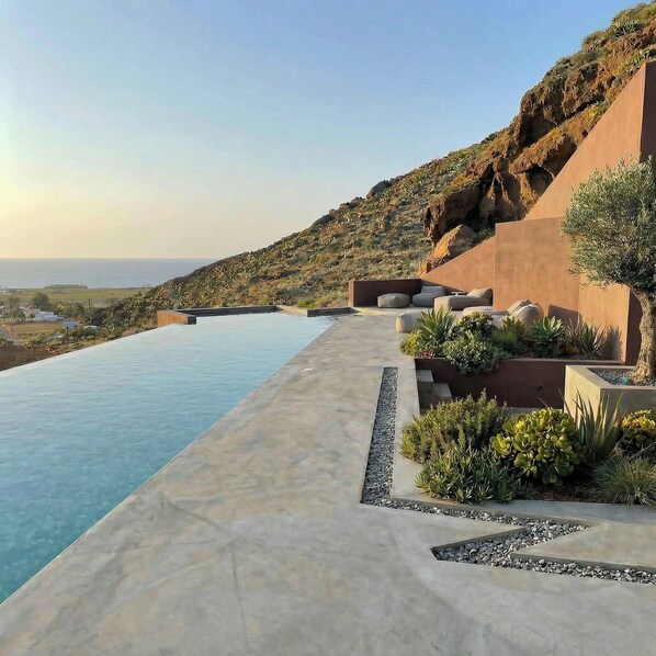 Ein moderner Infinity-Pool mit freiem Blick auf den Ozean, eine angelegte Grünfläche mit Pflanzen und ein Hügel im Hintergrund.