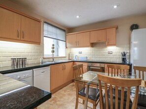 A kitchen with wooden cabinets, a glass dining table, and a built-in dishwasher. - 2 St. Catherines Cottages (Perth)