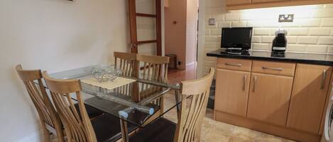 Un'area pranzo con un tavolo in vetro e sedie di legno, una cucina con mobili in legno e un piano di lavoro nero, e una porta che conduce in un'altra stanza.