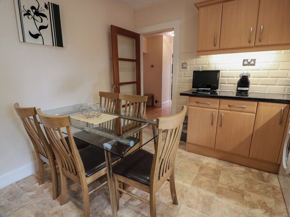 Un comedor con mesa de vidrio y sillas de madera, una cocina con armarios de madera y encimera negra, y una puerta que da a otra habitación.