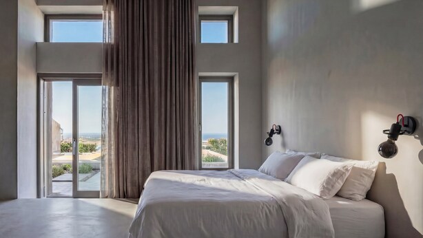 A bedroom with a large bed, two windows, a view of the outdoors, and a wall-mounted light fixture. - Dreamscape Villa, Anzo Villas (Thira - Santorini)