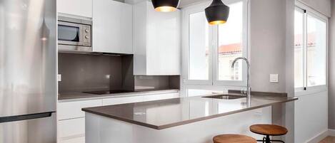 A modern kitchen with a stainless steel refrigerator, a built-in oven, a large island with stools, pendant lighting, and a window with a view.