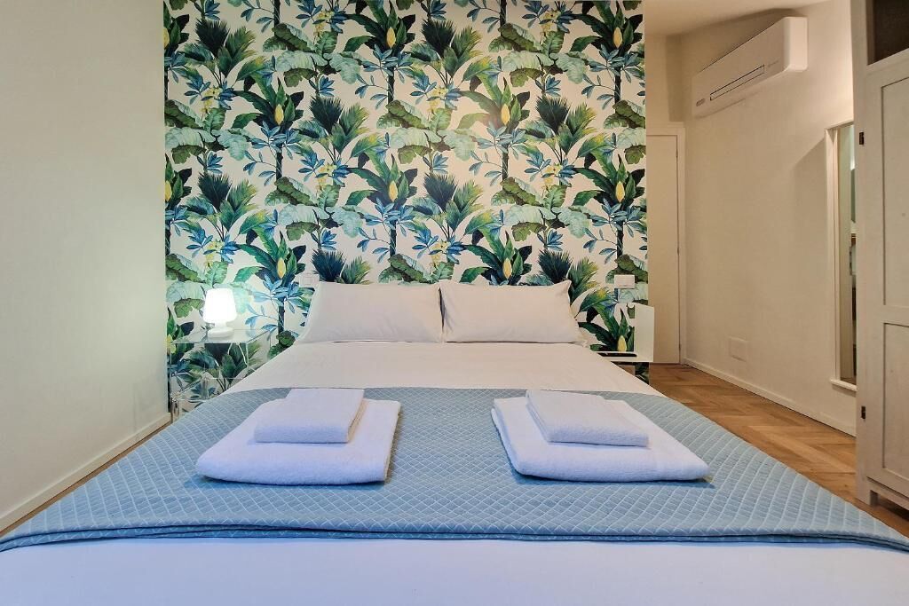 A hotel room with a bed, two pillows, a blue bedspread, and a floral wallpaper.