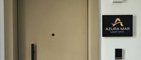 Una puerta cerrada con picaporte, un felpudo negro y un letrero en la pared que indica 'AZURA MAR LUXURY SUITE'.