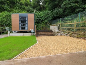 A modern building with a stone base and wooden upper structure, surrounded by a gravel area and a fenced garden. - Cwtch (Kidwelly)