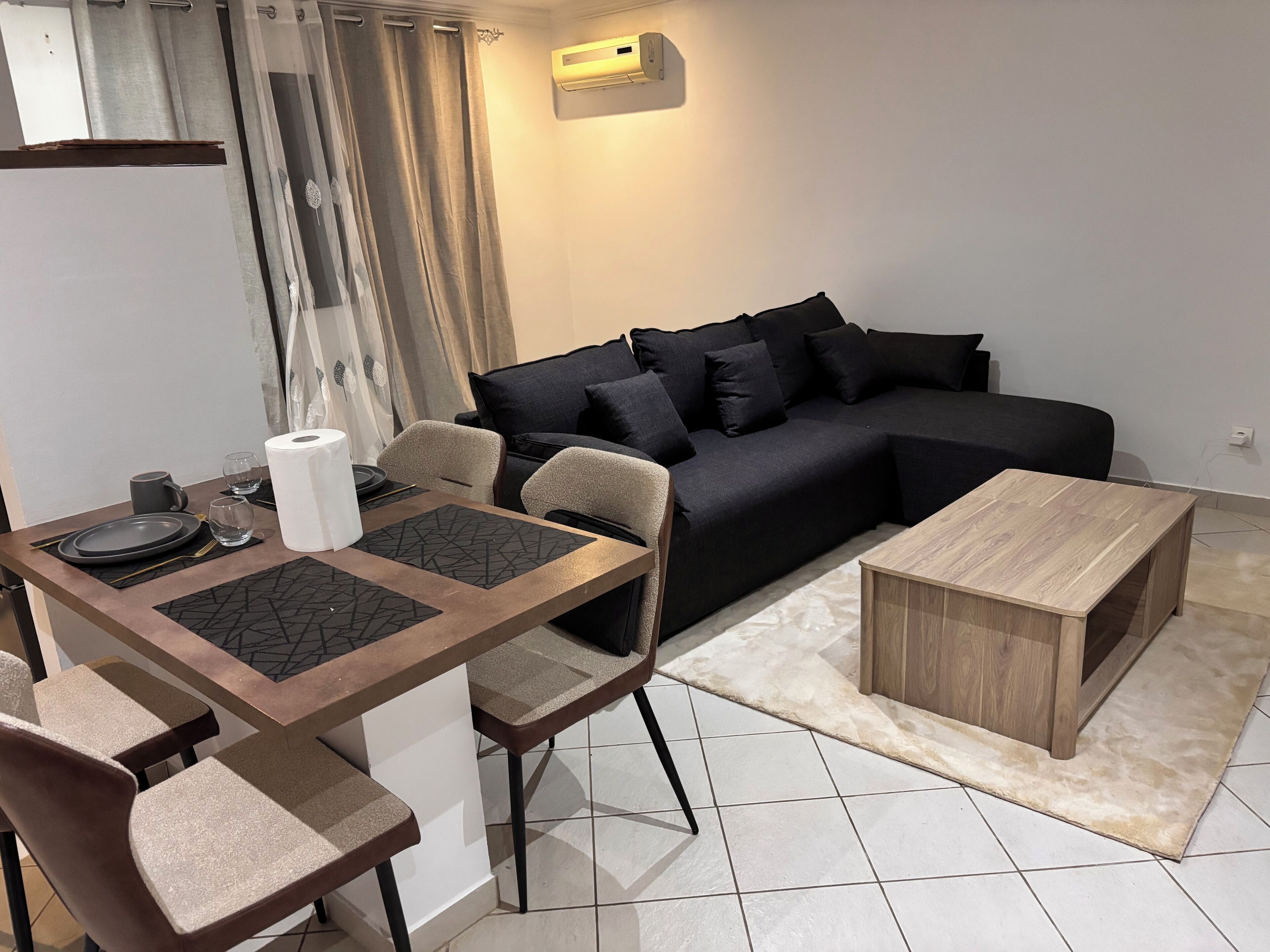 Un salon moderne avec un canapé noir, une table basse en bois et un coin repas avec une table dressée pour deux personnes.