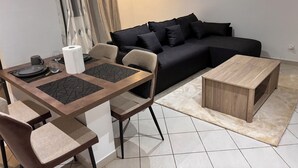 A modern living room with a black sofa, a wooden coffee table, and a dining area with a table set for two. - Luxury Apartment with Pool in Saly – Quiet and Secure Residential Complex (Saly)