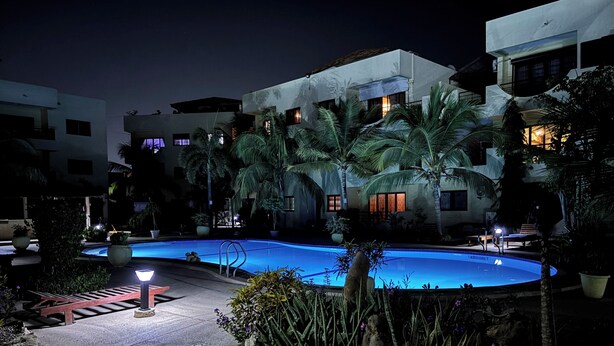 A nighttime swimming pool area with a pool, palm trees, and a building with illuminated windows. - Luxury Apartment with Pool in Saly – Quiet and Secure Residential Complex (Saly)