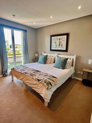 A bedroom with a bed, a nightstand, a lamp, a framed picture on the wall, and a view through the window. - Luxurious Secure 2 Bedroom V&A Waterfront Apt (Cape Town)