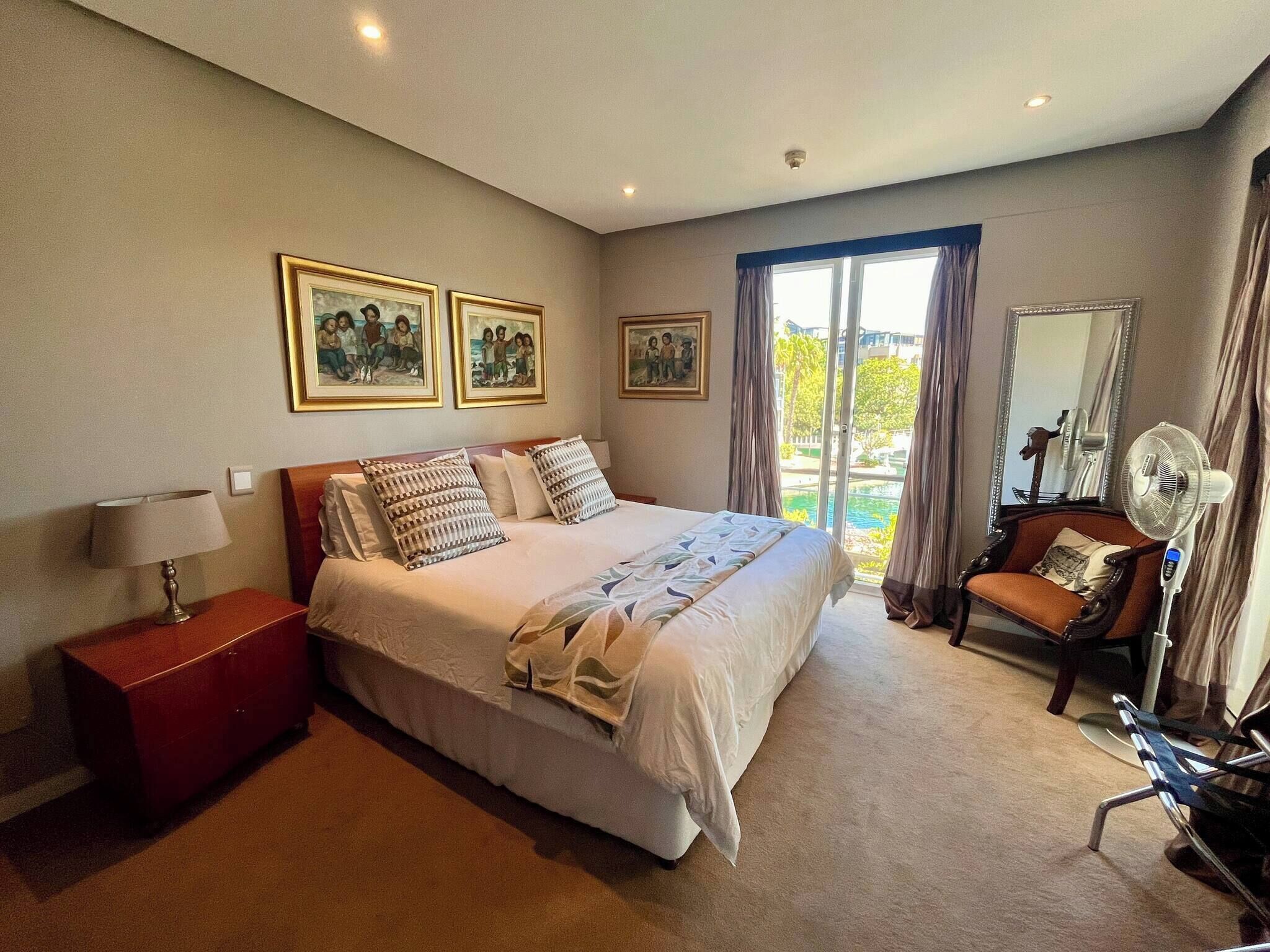 A bedroom with a bed, a nightstand, a chair, a fan, and a view of the outdoors.