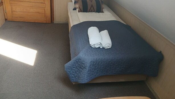 A single bed with a blue quilt and two rolled towels, a plaid pillow, and a wooden door in a room with a carpeted floor.