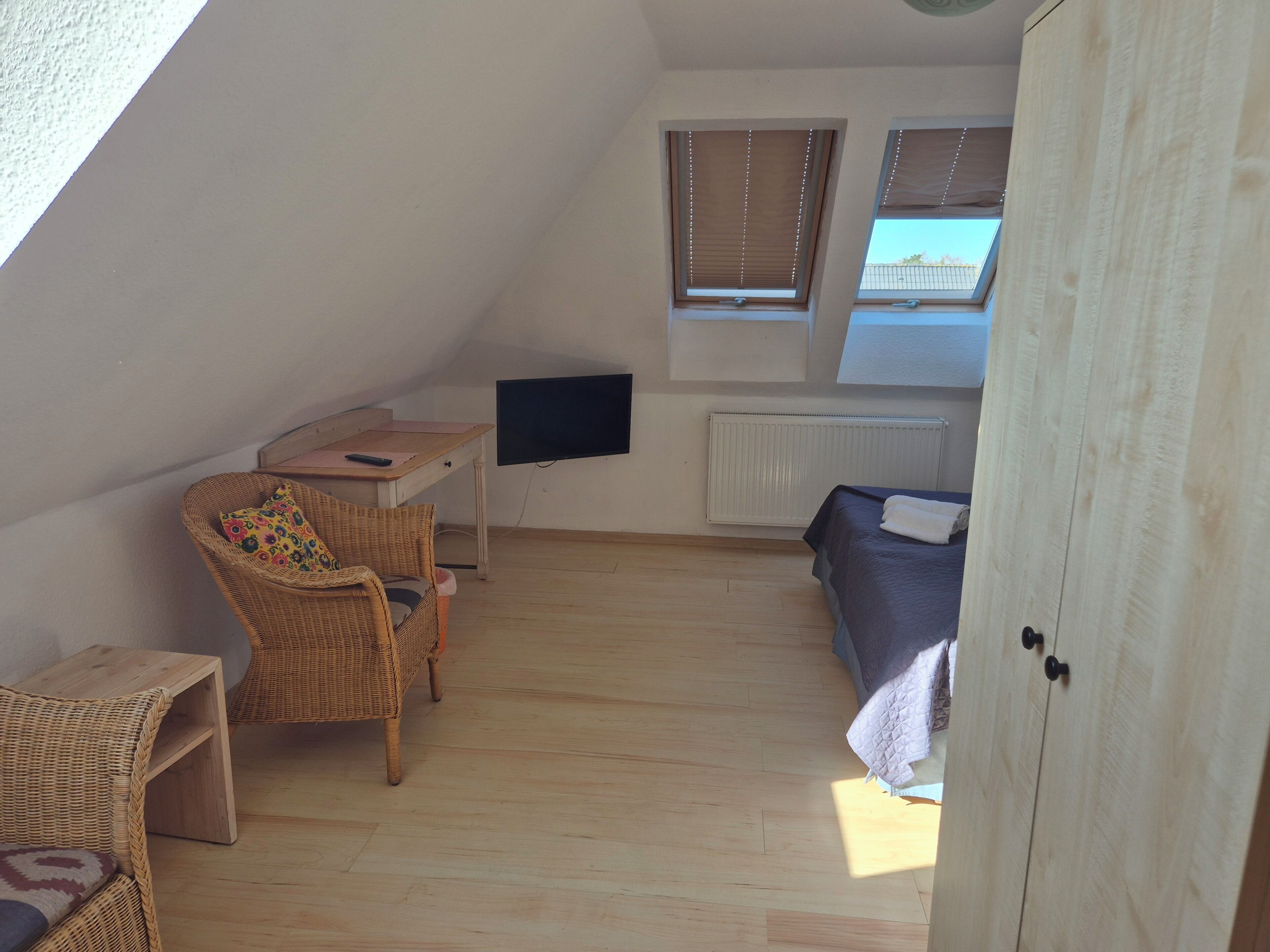 A small, well-lit room with a sloped ceiling, a bed, a desk with a chair, a television, and a window with blinds.