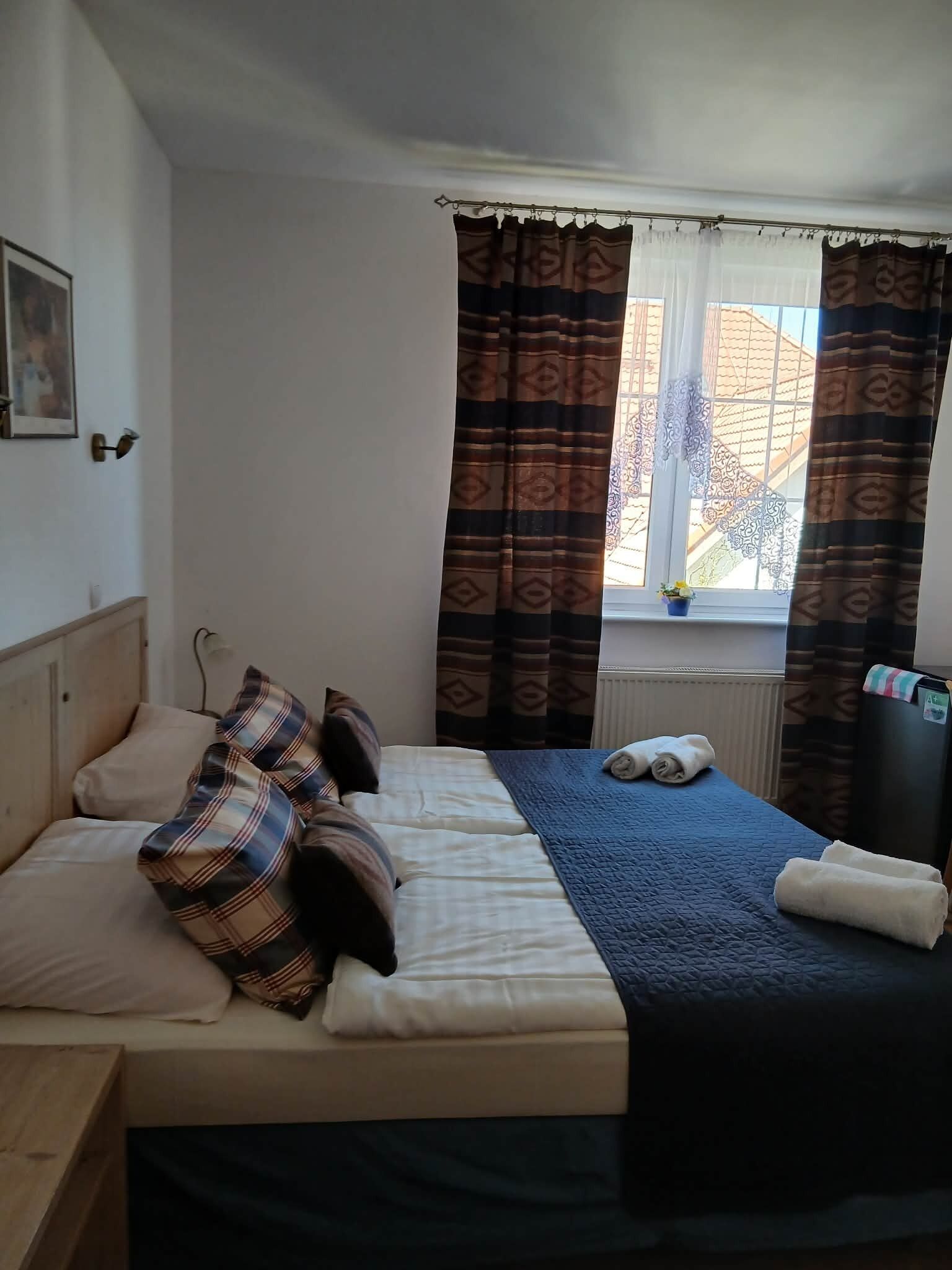Quarto com cama, almofadas, manta, janela com cortinas, radiador e uma pequena planta.