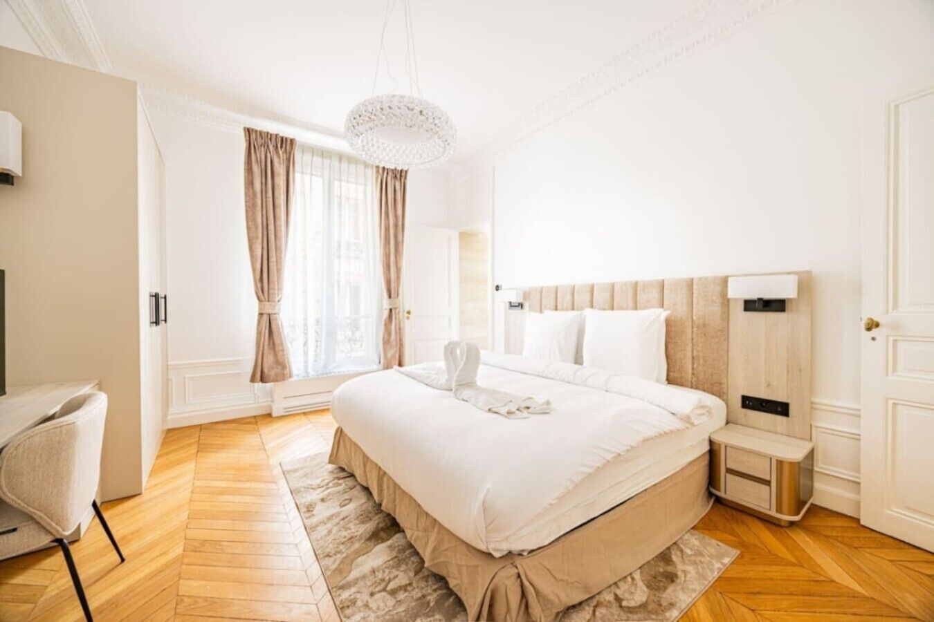 Una habitación de hotel con una cama grande, una mesita de noche, una silla, un ventanal con cortinas y un candelabro.